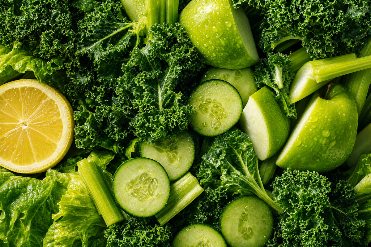Tightly packed raw green vegetables and fruit (kale, cucumber, apple, celery and lemon) filling the frame.