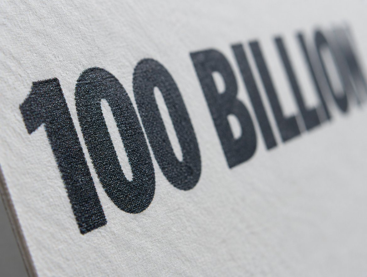 A close macro of the words 100 BILLION printed in heavy type on plain white carton board, near enough that the halftone dots of the ink and the fibre of the board are visible.